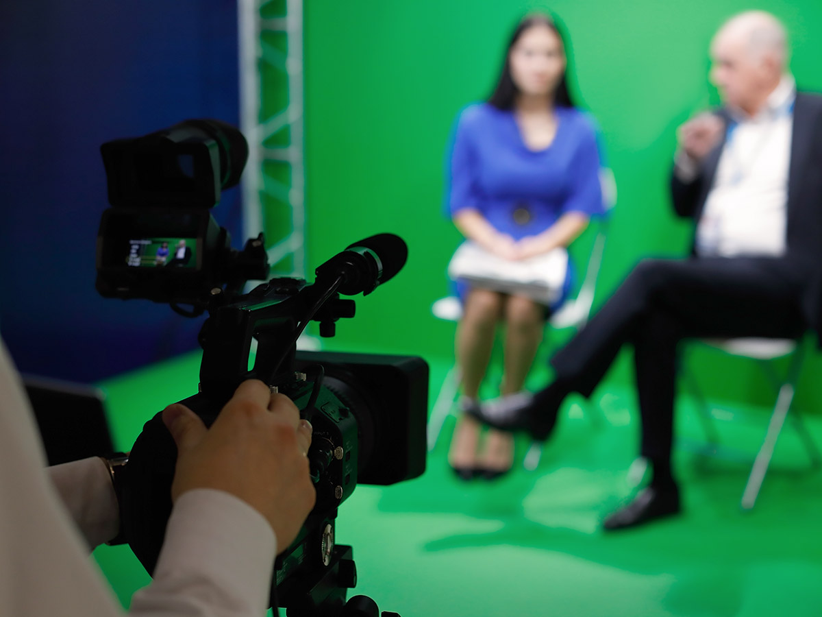 behind the video camera view of woman and man sitting in front of green screen